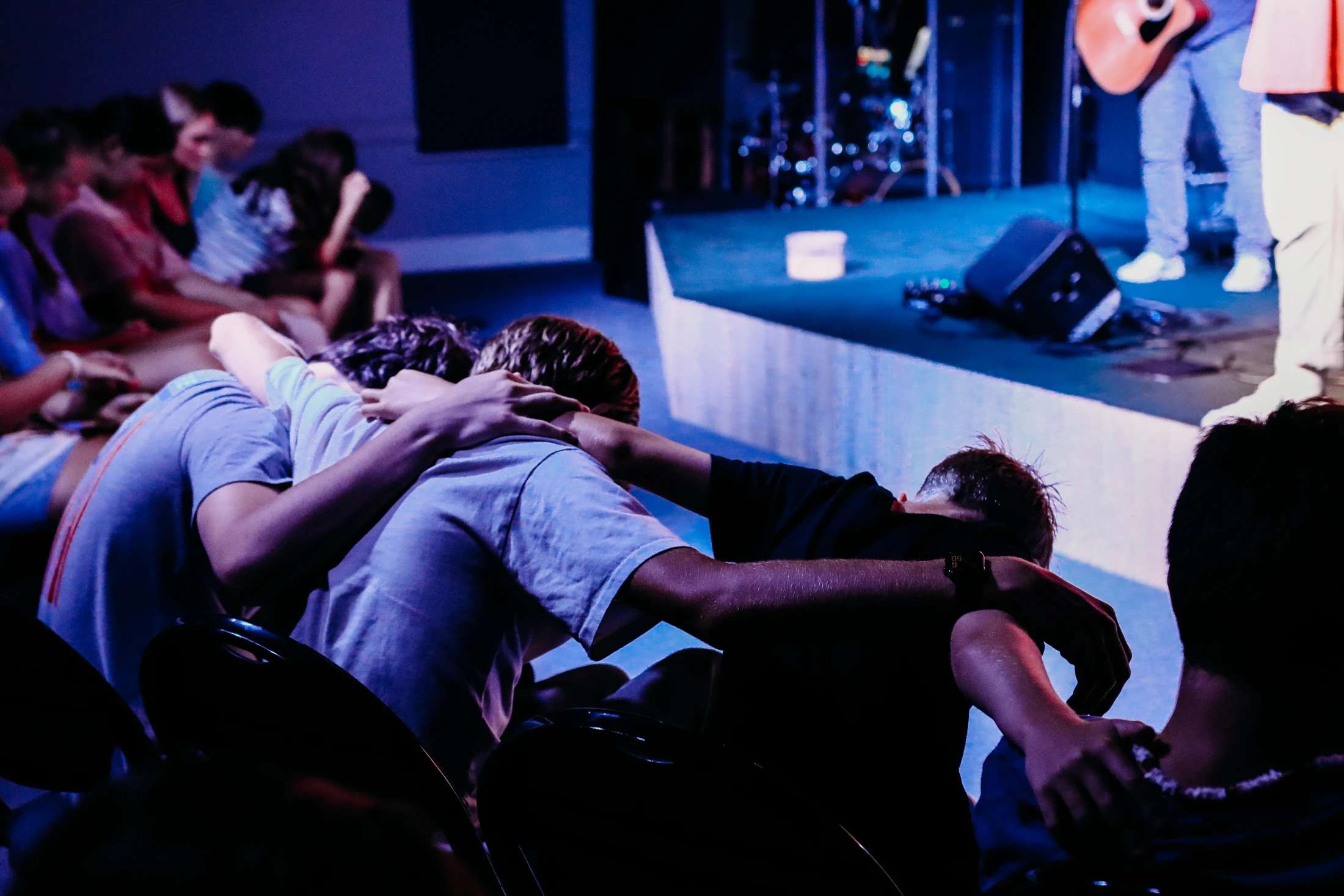 A group of young people huddle together with their arms around each other in a dimly lit room, facing a stage with a band performing under blue lights.